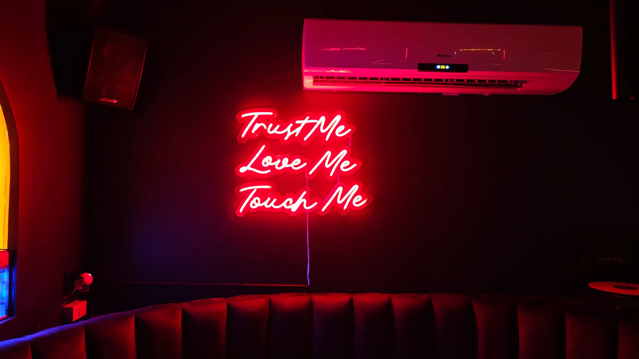 Red neon sign reading Trust Me Love Me Touch Me above a velvet banquette at a cocktail bar in Vientiane