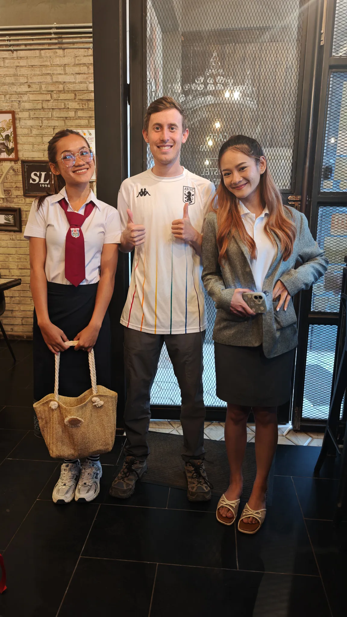 Full-length goodbye photo with two Lao friends at a cafe doorway in Vientiane wearing an Aston Villa shirt