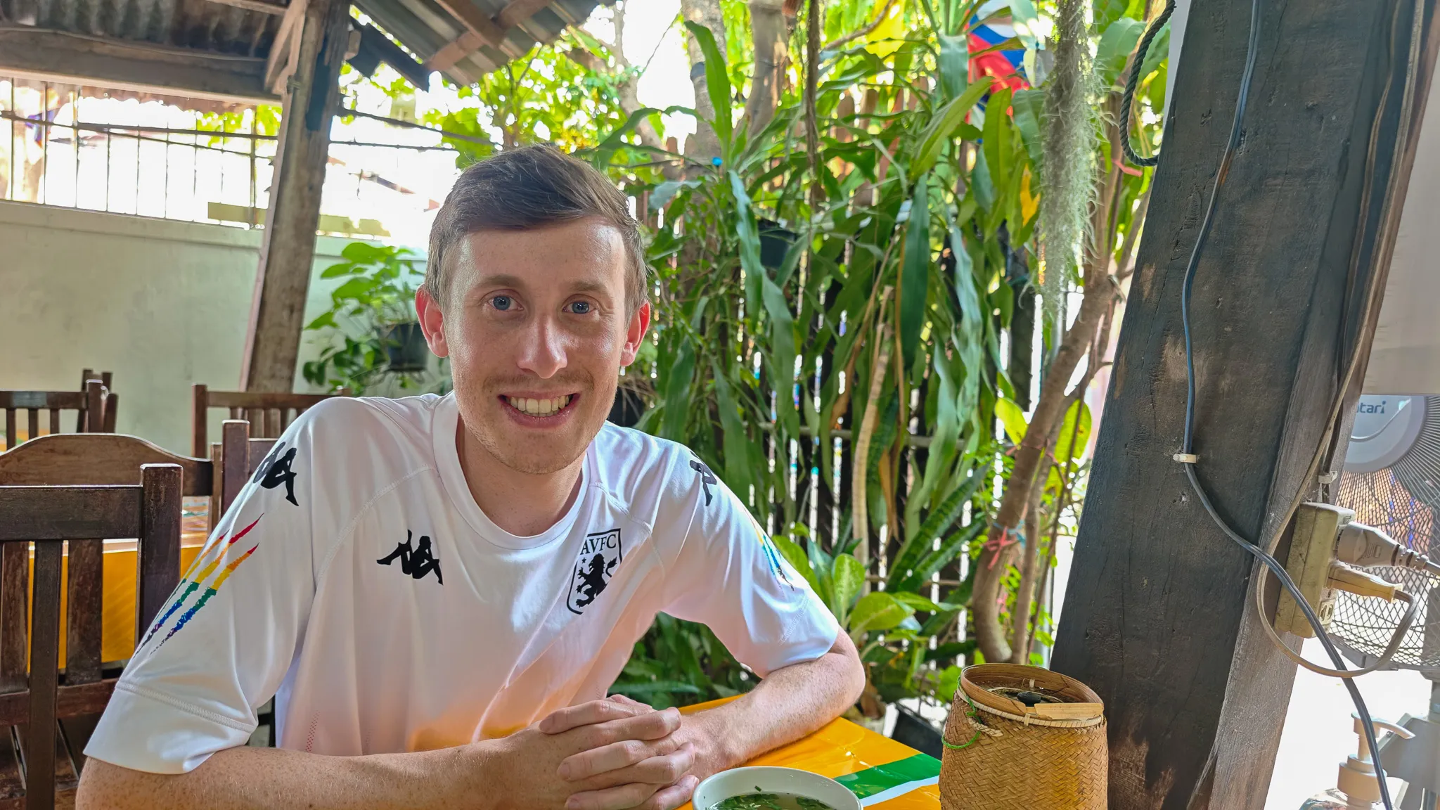 Smiling at a table with Lao food and sticky rice basket at Mark Wiens recommended restaurant in Vientiane