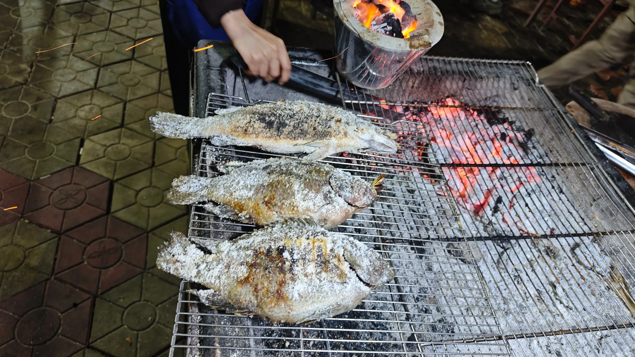 Salt-crusted whole fish being grilled over charcoal at a Mekong riverside restaurant in Vientiane
