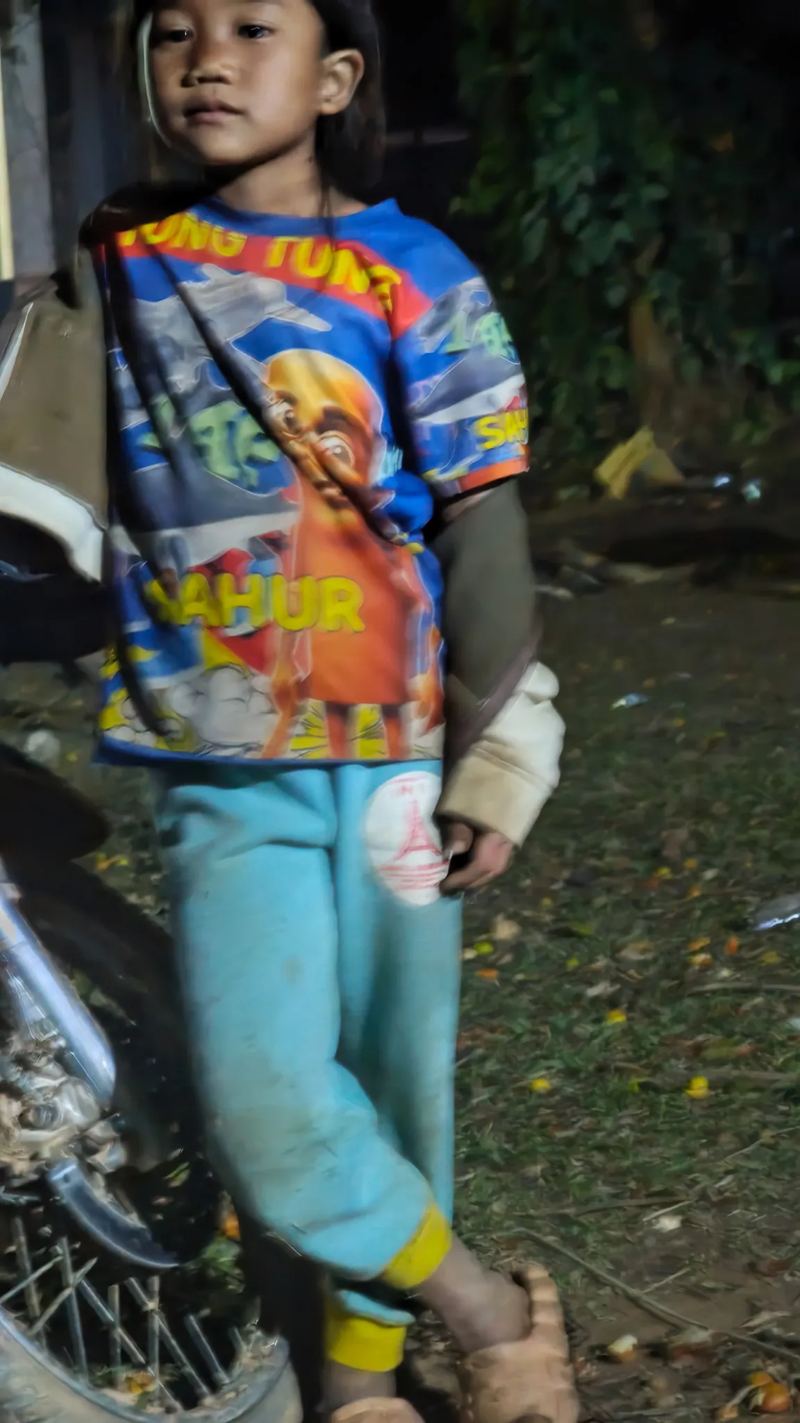 Young village child standing by a motorbike at night in Ban Haddean, wearing a colourful cartoon T-shirt