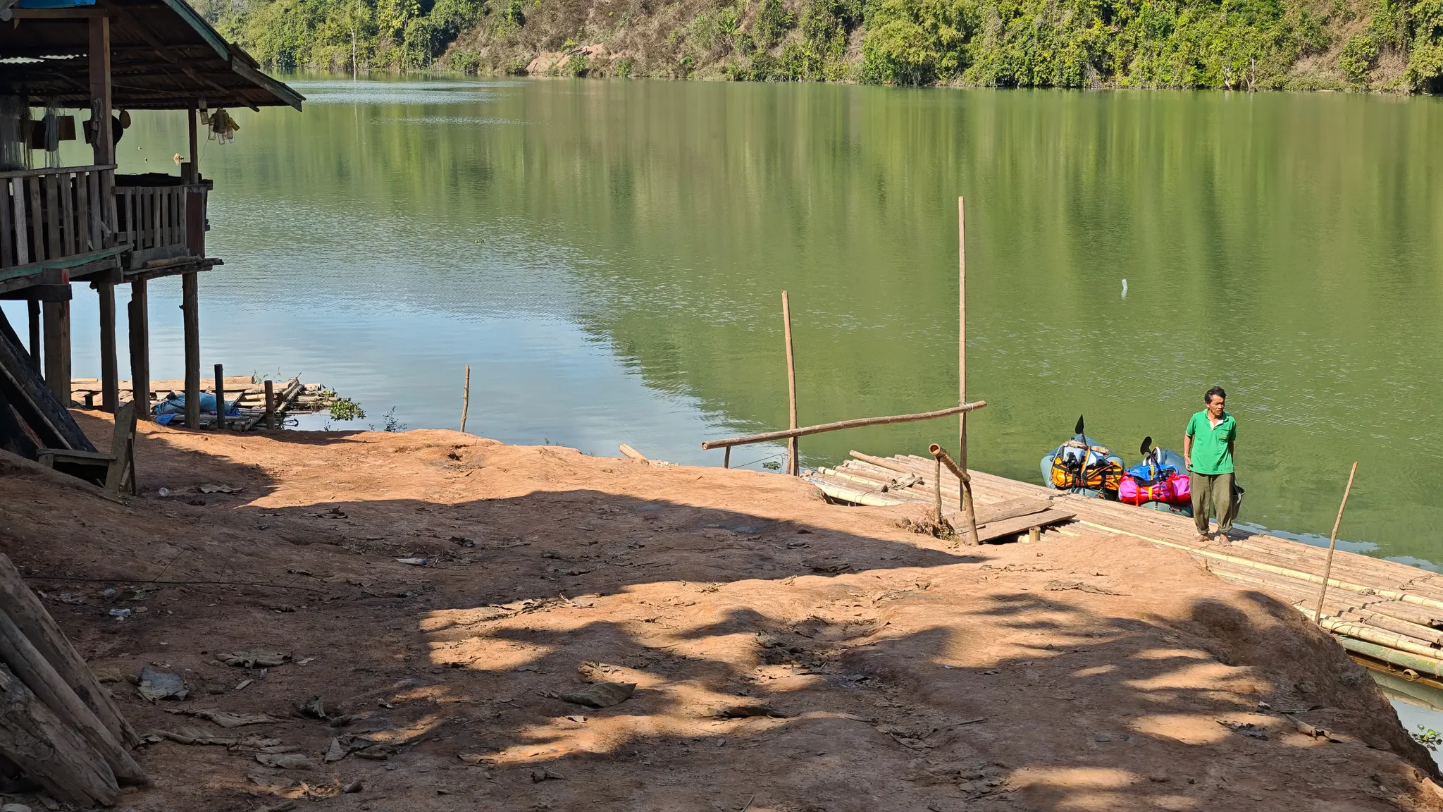 A stilted village hut and bamboo landing with packrafting gear laid out beside the calm green Nam Ou river
